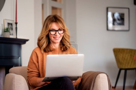 Foto de An attractive middle aged woman sitting in the armchair at home and using laptop. Beautiful female wearing casual clothes while working from home. Home office. - Imagen libre de derechos