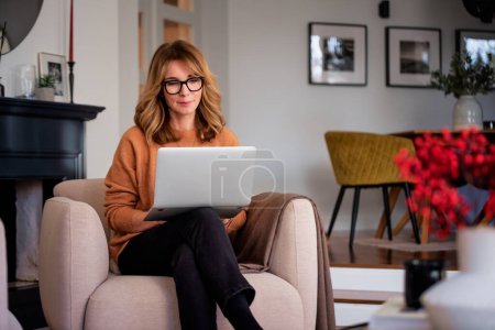 Téléchargez les photos : An attractive middle aged woman sitting in the armchair at home and using laptop. Beautiful female wearing casual clothes while working from home. Home office. - en image libre de droit