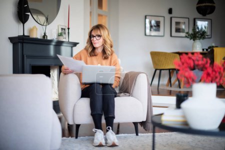 Foto de Mujer de mediana edad usando un portátil y sosteniendo el documento en su mano mientras está sentada en casa y trabajando. Oficina en casa. - Imagen libre de derechos
