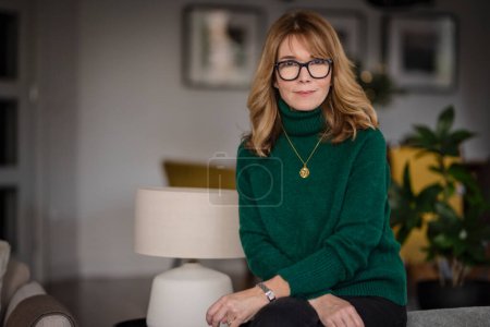 Foto de Portrait of attractive middle aged woman relaxing in an armchair at home. Blond haired female wearing eyeglasses and sweater. - Imagen libre de derechos