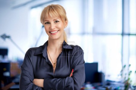 Téléchargez les photos : Heureuse femme d'affaires aux cheveux blonds debout au bureau avec les bras croisés. - en image libre de droit