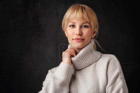 Photo for Beautiful smiling woman standing at isolated dark background. Blond haired woman wearing turtleneck sweater. Copy space. - Royalty Free Image