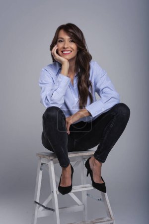 Photo for Full length shot of attractive brunette woman wearing shirt and black trousers while sitting on stool at isolated grey background. Copy space. - Royalty Free Image