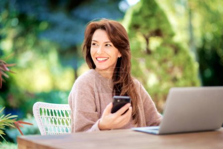 Foto de Mensajería de texto mujer de mediana edad de confianza y el uso de la computadora portátil mientras está sentado en el balcón. Atractiva hembra con suéter y sonrisa alegre. - Imagen libre de derechos