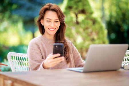 Photo for Confident middle aged woman text messaging and using laptop while sitting on balcony. Attractive female wearing sweater and cheerful smiling. - Royalty Free Image