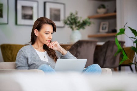 Foto de Shot of an attractive middle aged woman sitting in an armchair while using laptop at home. Blank screen. Beautiful female looking thoughtfully and wearing casual clothes. - Imagen libre de derechos