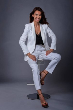 Téléchargez les photos : Plan studio complet d'une jolie femme d'âge moyen portant un costume blanc et riant assis sur un fond gris isolé. Espace de copie. - en image libre de droit