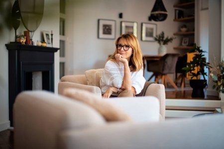 Foto de Retrato de una atractiva mujer de mediana edad que se relaja en un sillón en casa. Mujer de pelo rubio con gafas y camisa blanca y soñando despierto. - Imagen libre de derechos