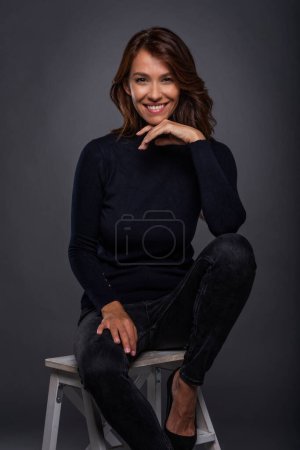 Foto de Attractive middle aged woman with toothy smile wearing turtleneck sweater while sitting on stool against at isolated dark background. Copy space. Studio shot. - Imagen libre de derechos