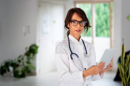 Photo for Female doctor wearing lab coat and stethoscope and using digital tablet for work while standing at hospital corridor. Smiling healthcare worker looking at camera and smiling. - Royalty Free Image