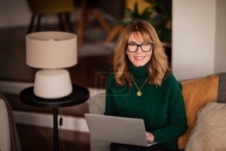 Téléchargez les photos : Femme d'âge moyen souriante avec ordinateur portable assis à la maison et au travail. Bureau à domicile. - en image libre de droit