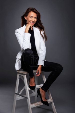 Téléchargez les photos : Full length of attractive middle aged woman with toothy smile wearing white blazer while sitting on stool against at isolated dark background. Copy space. Studio shot. - en image libre de droit