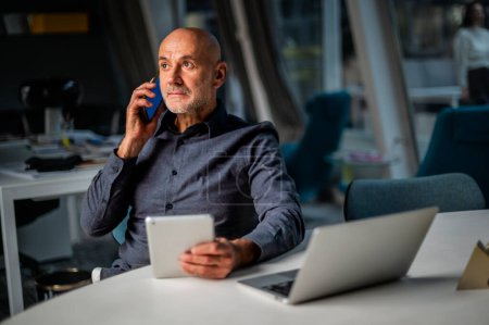 Foto de Empresario usando tableta diigital y haciendo una llamada mientras está sentado en una oficina moderna. Multitarea. - Imagen libre de derechos