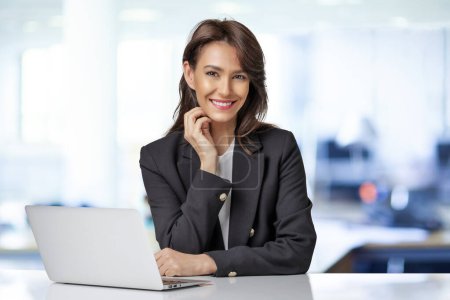 Photo for Attractive businesswoman wearing blazer and using laptop for work. Smiling female looking at camera and smiling. - Royalty Free Image