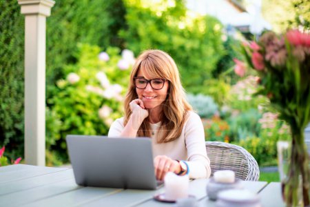 Photo for Attractive mid aged woman using her laptop for video conferences while sitting at backyard at home. Home office. - Royalty Free Image