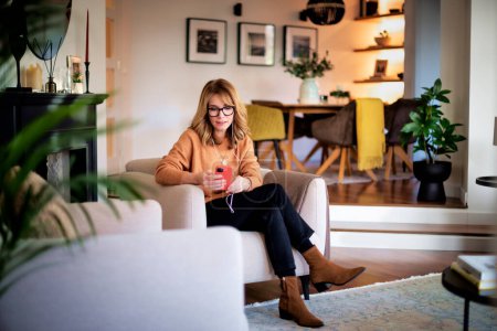 Foto de Longitud completa de una mujer sentada en casa y usando auriculares y teléfonos inteligentes mientras recibe videollamadas. - Imagen libre de derechos