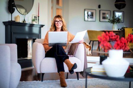 Foto de Mujer de mediana edad usando un portátil y sosteniendo el documento en su mano mientras está sentada en casa y trabajando. Oficina en casa. - Imagen libre de derechos