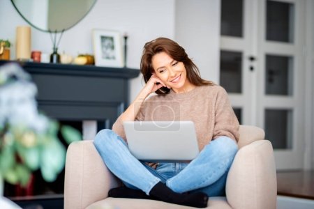 Shot of an attractive middle aged woman sitting in an armchair while using laptop and having video call. Beautiful female wearing casual clothes. Poster 644556478