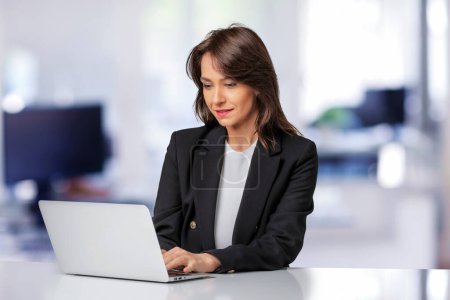 Téléchargez les photos : Femme d'affaires attrayante portant un blazer et utilisant un ordinateur portable pour le travail. Femme souriante assise au bureau et travaillant. - en image libre de droit