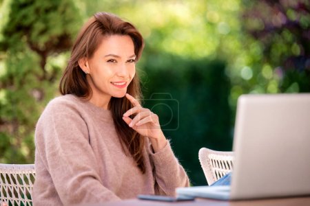 Téléchargez les photos : Femme d'âge moyen confiante utilisant un ordinateur portable tout en étant assis sur le balcon à la maison. Attrayant pull féminin et souriant gai. - en image libre de droit
