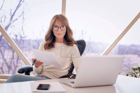Téléchargez les photos : Prise de vue d'une femme d'affaires mature assise au bureau et faisant de la paperasse tout en travaillant sur un ordinateur portable au bureau. - en image libre de droit