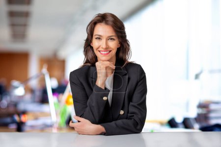 Foto de Atractiva mujer de negocios vistiendo chaqueta negra mientras está sentada en la oficina. Copiar espacio. Mano en la barbilla. - Imagen libre de derechos