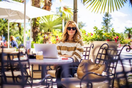 Téléchargez les photos : Prise de vue d'une heureuse femme d'âge moyen assise dans un café extérieur. Homme d'affaires confiant portant des vêtements décontractés et utilisant un ordinateur portable tout en travaillant en ligne. - en image libre de droit
