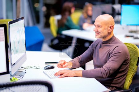 Foto de Sonriente hombre de negocios de mediana edad sentado en el escritorio de la oficina y usando computadoras para el trabajo. - Imagen libre de derechos