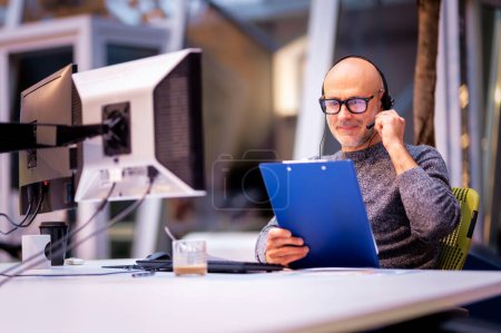 Mann mittleren Alters mit Kopfhörer auf Videokonferenz im modernen Büro. Professionelle männliche mit Computern und Klemmbrett umgeben, während sie am Schreibtisch sitzen und arbeiten. 