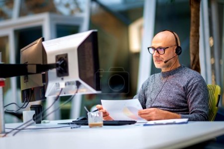 Téléchargez les photos : Homme d'affaires mature avec écouteurs assis à la table et travaillant sur des ordinateurs. Assistant de service client masculin. - en image libre de droit