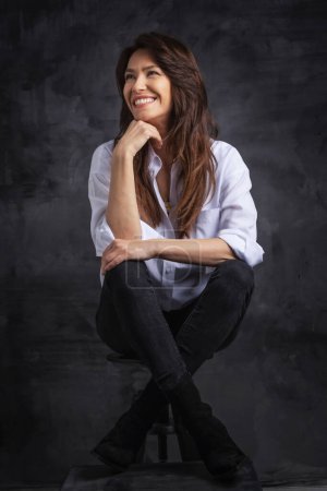 Foto de Atractiva mujer de mediana edad con sonrisa dentada que usa camisa blanca y jeans negros mientras está sentada en el taburete contra un fondo oscuro aislado. Copiar espacio. Captura de estudio. - Imagen libre de derechos