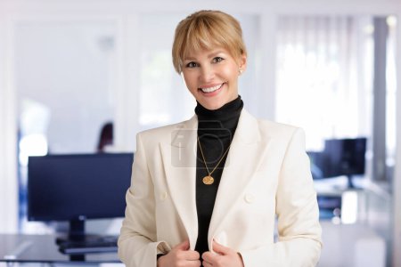 Photo for Close-up of businesswoman smiling and looking at camera. Blond haired female wearing white blazer while standing at the office. Copy space. - Royalty Free Image