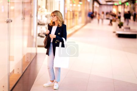 Foto de Longitud completa de una atractiva mujer madura con bolsa de compras en el centro comercial de la ciudad. Mujer de pelo rubio con blazer y jeans. - Imagen libre de derechos