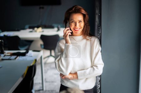 Photo for Attractive business woman using mobile phone and having a call while standing in a modern office. - Royalty Free Image