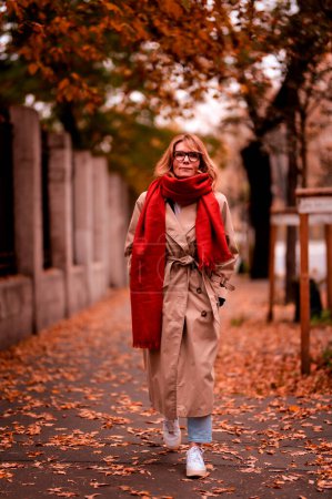 Photo for Fashionable middle aged woman walking in the street and enjoying autumn day. Attractive female wearing scarf and trench coat outdoor. - Royalty Free Image