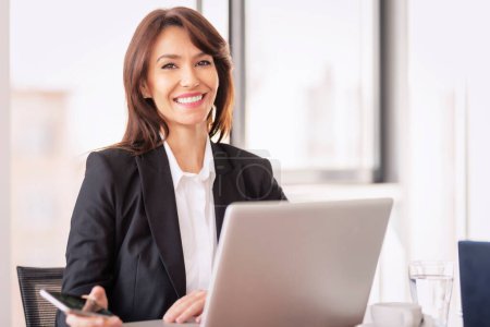 Foto de Atractiva mujer de negocios con chaqueta y el uso de la computadora portátil para el trabajo. Mujer confiada mirando a la cámara y sonriendo alegre. - Imagen libre de derechos