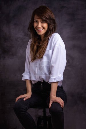 Foto de Retrato de estudio de una mujer atractiva que usa camisa blanca y jeans mientras está sentada en un fondo oscuro aislado. Morena de mediana edad pelo femenino alegre sonriente. Copiar espacio. - Imagen libre de derechos