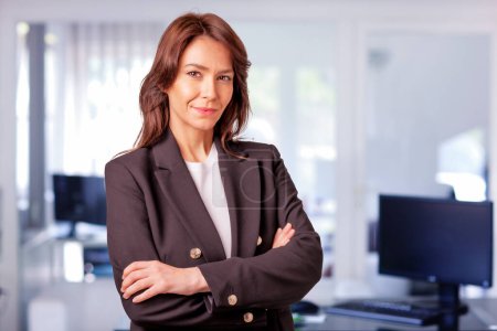 Photo for Attractive mid aged businesswoman wearing blazer and standing at the office. Confident professional woman looking at camera and smiling. - Royalty Free Image