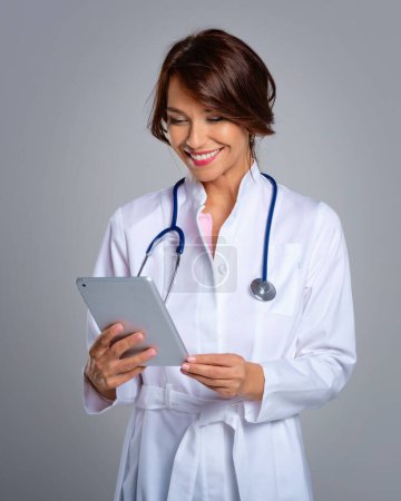 Photo for Portrait of a female doctor holding her patient chart on digital tablet while standing against light grey background. - Royalty Free Image