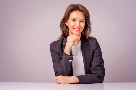 Photo for Portait of an attractive businesswoman wearing black blazer while sitting at isolated grey background. Copy space. Hand on chin. - Royalty Free Image