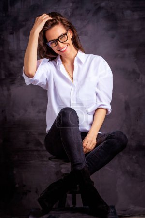 Foto de Atractiva mujer de mediana edad con sonrisa dentada usando camisa blanca y jeans negros mientras se sienta en un fondo oscuro aislado. Copiar espacio. Grabado en estudio. Longitud completa. - Imagen libre de derechos