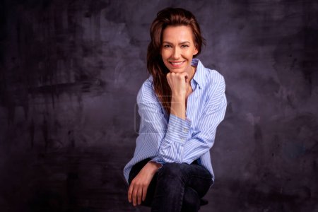 Foto de Atractiva mujer de mediana edad con sonrisa dentada usando camisa azul y jeans negros mientras se sienta en un fondo oscuro aislado. Copiar espacio. Captura de estudio. - Imagen libre de derechos