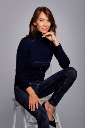Photo for Shot of attractive brunette haired woman sitting on stool at isolated grey background. Copy space. - Royalty Free Image