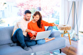 Happy couple relaxing on the couch together. Cheerful woman and handsome man using laptop while browsing on the internet. Man showing something on the screen with his finger.  Poster #657161824