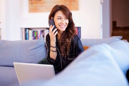 Photo for Attractive brunette haired woman cheerful smiling. Mid aged female using mobile phone and laptop for work. Home office. - Royalty Free Image