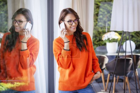 Téléchargez les photos : Portrait d'une femme joyeuse faisant un appel téléphonique alors qu'elle se tenait à la fenêtre à la maison. Brunette aux cheveux féminins portant un pull orange et un jean bleu. - en image libre de droit