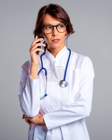 Téléchargez les photos : Portrait d'une femme médecin pensante utilisant un téléphone portable et parlant. Fond gris isolé. Un travailleur de la santé confiant porte une blouse de laboratoire. - en image libre de droit