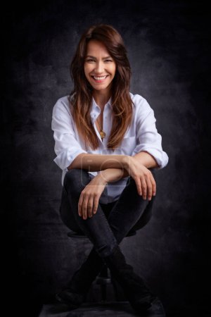 Foto de Atractiva mujer de mediana edad con sonrisa dentada usando camisa blanca y jeans negros mientras se sienta en un fondo oscuro aislado. Copiar espacio. Captura de estudio. - Imagen libre de derechos