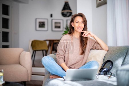 Photo for Shot of an attractive middle aged woman sitting on the sofa while using laptop. Beautiful female wearing casual clothes and looking thoughtfully. - Royalty Free Image