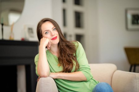 Photo for Portrait of beautiful long haired woman wearing casual clothes while relaxing in an armchair at home. Attractive young female wearing red lipstick and green sweater. - Royalty Free Image